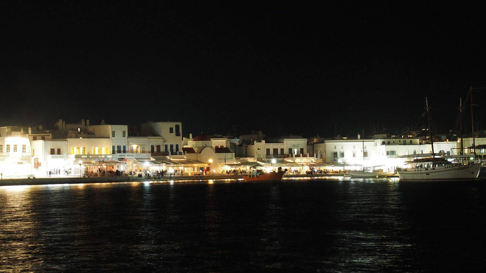 The lit waterfront of Mykonos Town at night, seen across the dark water of the harbour